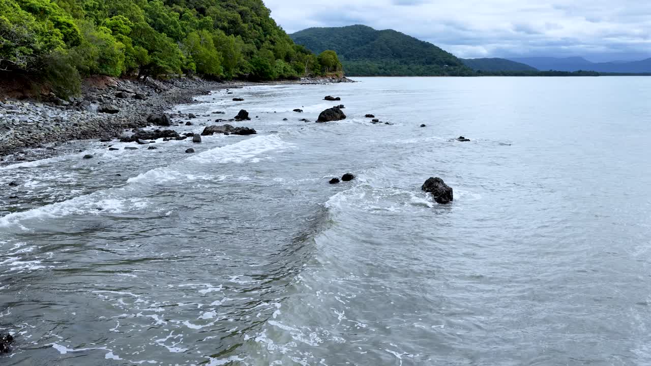 Camera slowly pans along rocky coastline, tropical forest, and calm ocean under overcast daylight