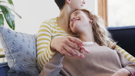 feliz pareja de lesbianas caucásicas sentadas, abrazadas y tomadas de la mano en el sofá en la casa soleada