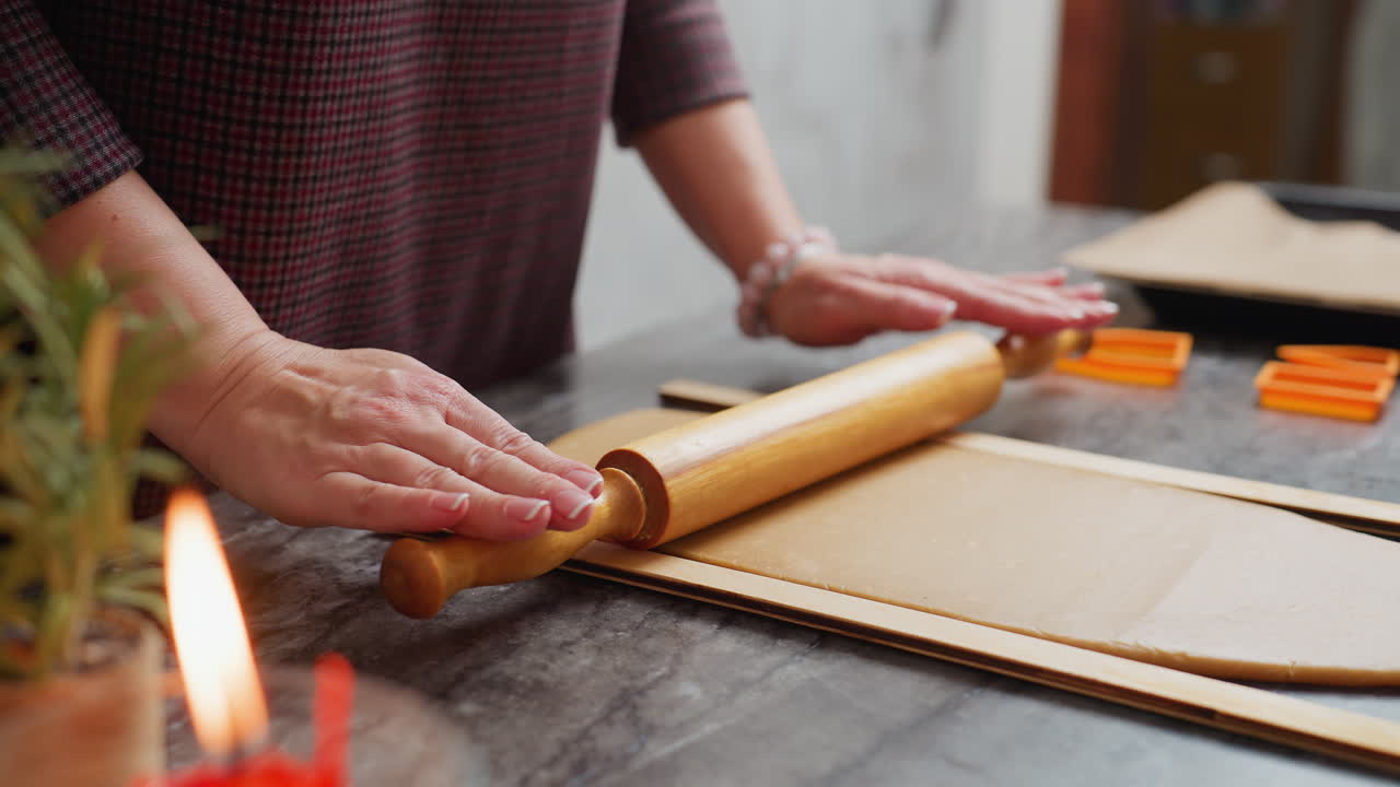 Close-up of lady hands rolling brown dough with wooden rolling pin, left wrist adorned with pink beaded bracelet, slight blur in background featuring candle, baking tools