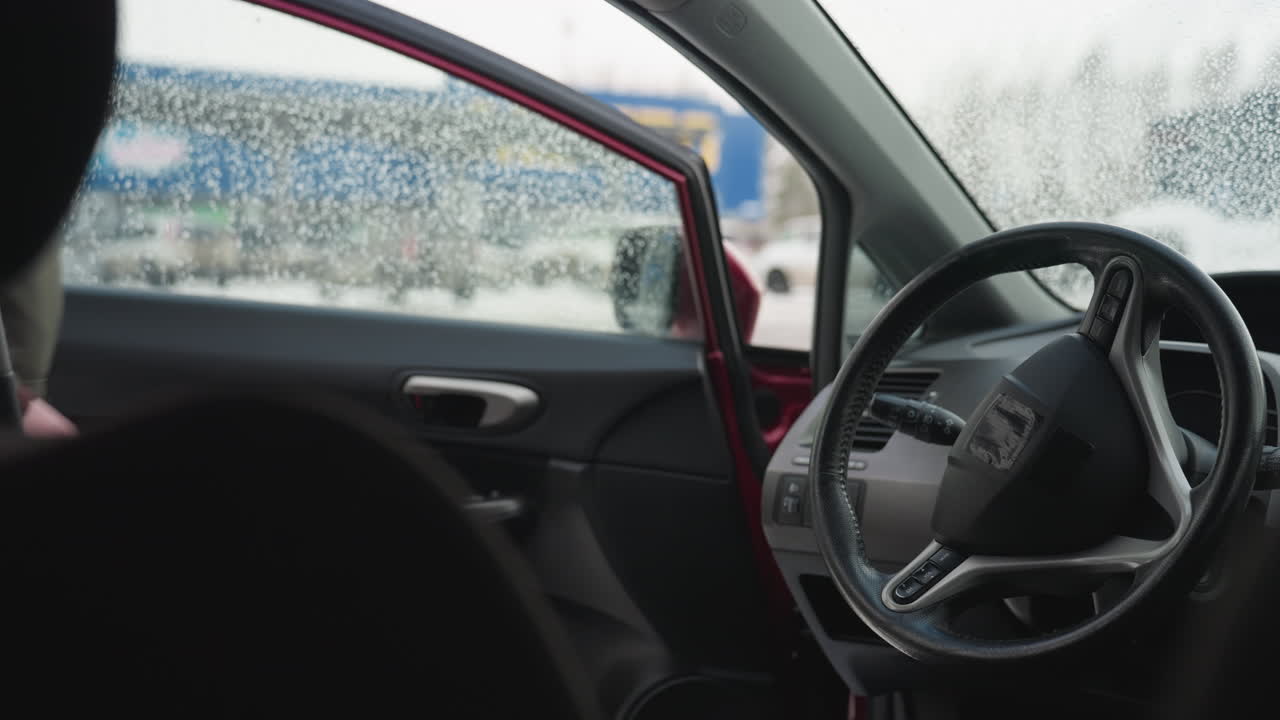 Close up of car interior showing steering wheel and wet window with water droplets, driver reflected in side mirror approaches car and opens door in cold snowy urban setting during winter