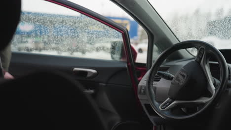 close up of car interior showing steering wheel and wet window with water droplets, driver reflected in side mirror approaches car and opens door in cold snowy urban setting during winter