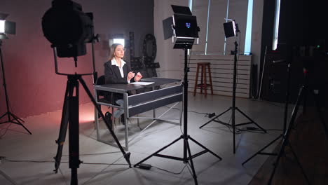 confident female blogger sitting at desk talks to camera. brunette in black jacket speaks and gestures. soffits light surround the blogger.