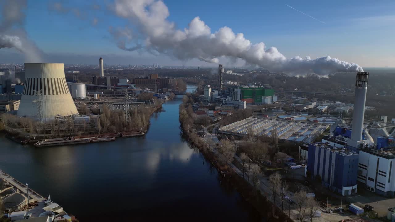 large industrial power plant emitting large amounts of smoke over the city Berlin next to a river on a sunny day. Spectacular aerial view flight descending drone
