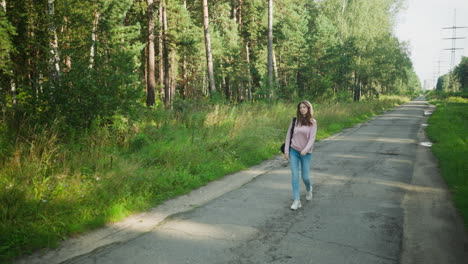 young student in pink hoodie and jeans walks calmly down empty cracked road surrounded by dense forest and tall grass, carrying backpack, enjoying peaceful nature on quiet sunny day