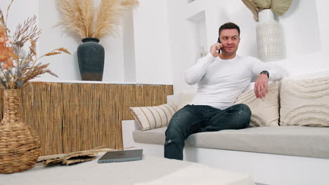 man sitting relaxed on the sofa speaks on the phone. man is smiling while talking to somebody.