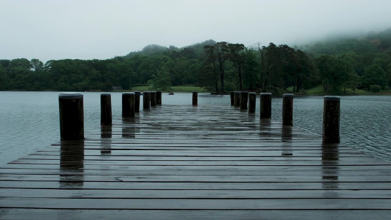 Inquadratura fissa di un pontile di legno bagnato dalla pioggia che si estende nel lago Coniston, con la nebbia che incombe sulla riva e sul bosco circostante nel Lake District.