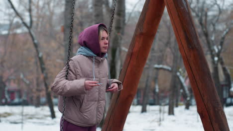 side view of young woman dressed for winter on swing holding chains, hoodie rope in maroon trousers, snowy background with trees, building, and moving car in the distance