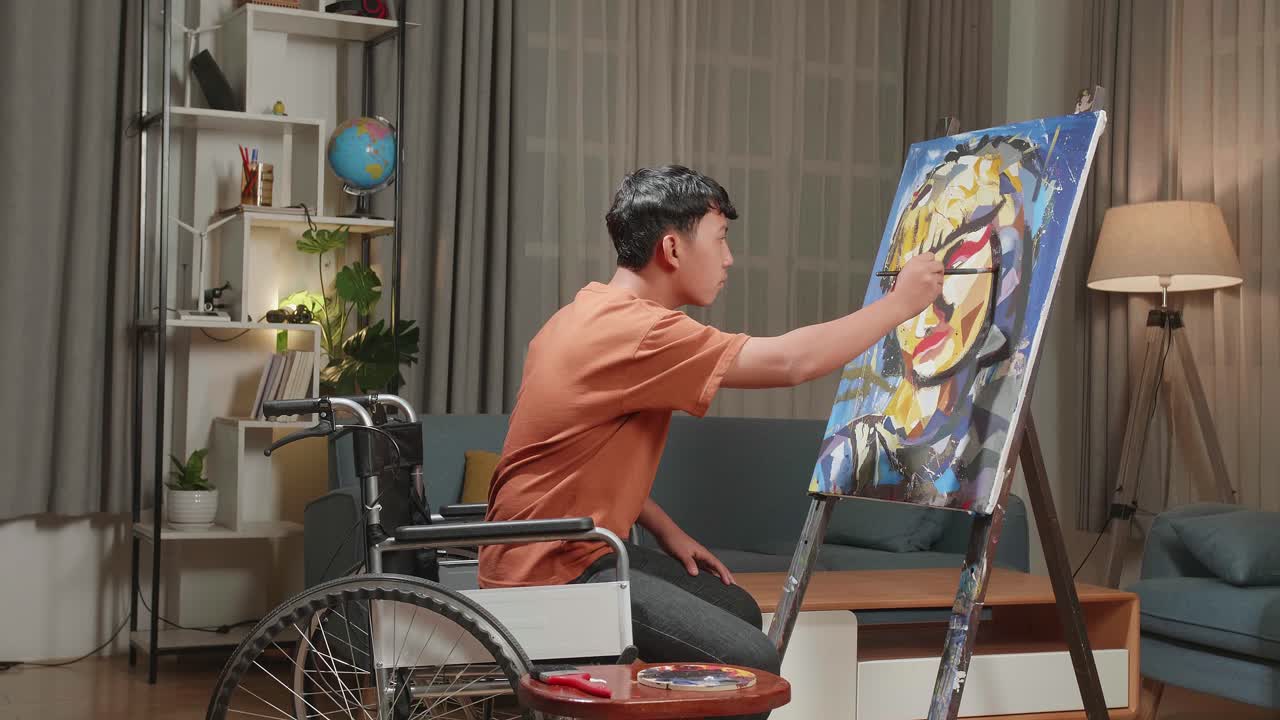 Hind View Side View Of An Asian Artist Boy In Wheelchair Holding Paintbrush Mixed Colour And Painting A Girl On The Canvas