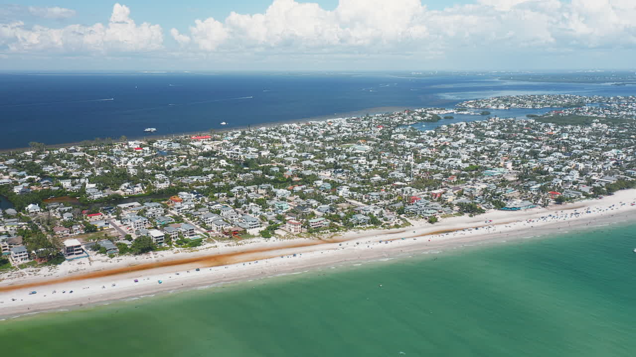 The coastline of Anna Maria Island features a wide sandy beach meeting turquoise waters, with a residential community stretching inland under a bright summer sky