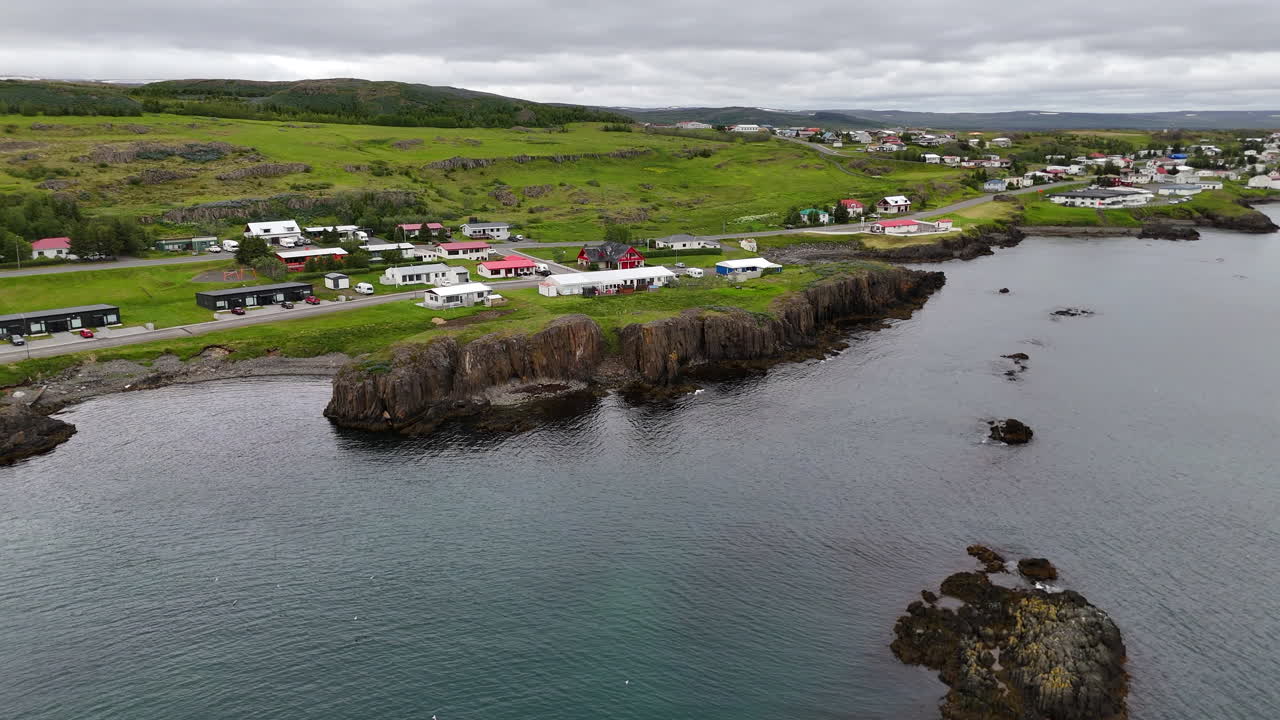 Aerial drone view of Vopnafjörður in Austurland, showing a dramatic coastline, dark cloudy skies, wide open fields, and the calm shoreline of this remote Icelandic region