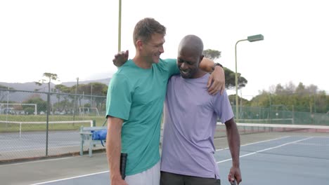 portrait of two happy diverse male friends embracing on outdoor tennis court after game, slow motion