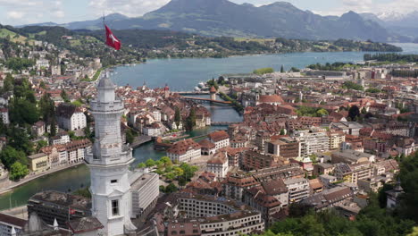 Drone-Volando-Más-Allá-De-La-Bandera-Suiza-En-La-Torre-Del-Castillo-Y-Revelando-La-Hermosa-Ciudad-De-Luzern,-Suiza