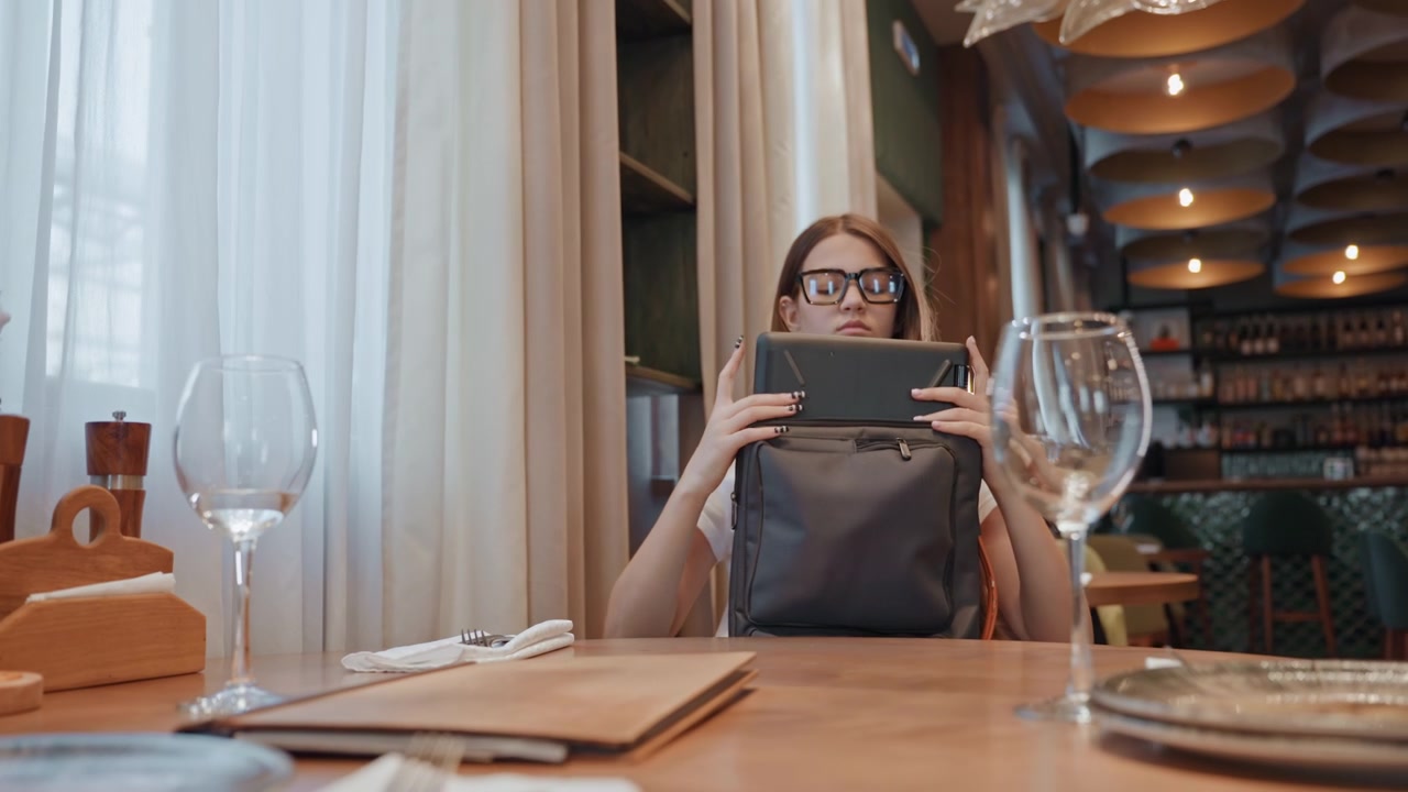Young woman checking laptop privacy in restaurant, lifting device from bag and scanning surroundings, wine glasses and place setting in foreground, ambient ceiling lights and modern interior, cautious