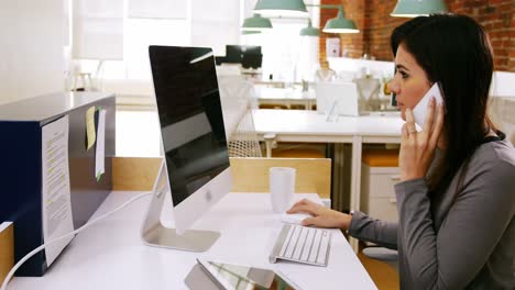 Female-executive-talking-on-mobile-phone-while-working-on-computer-at-desk