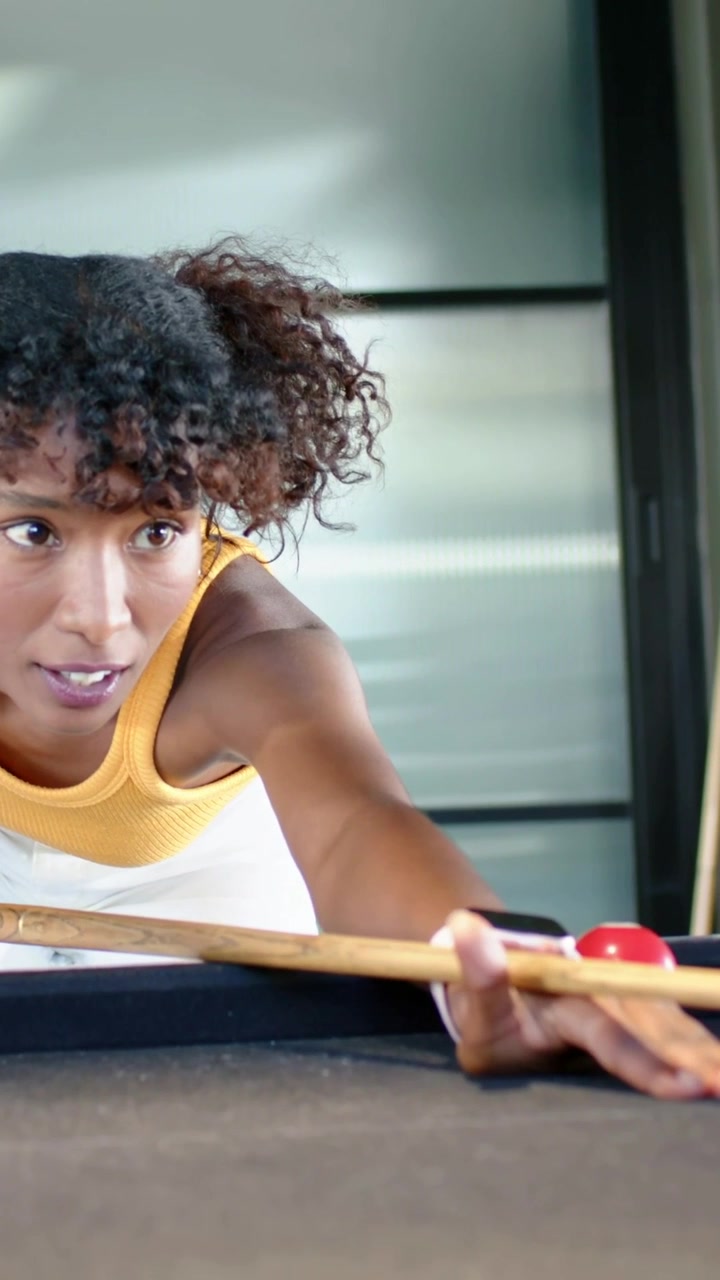 Vertical video: Approaching pool table, woman in yellow top lining shot, nudging red ball with cue