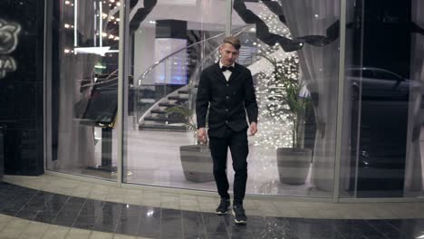 Attractive-young-man-in-a-black-suit-walking-out-of-a-hotel-in-the-evening.-Behind-a-lot-of-blurred-lights-and-glass-door-of-luxurious-hotel