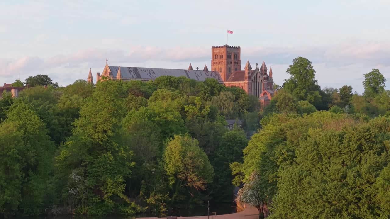 A cinematic drone glides behind the trees, revealing St Albans Park with its shimmering lake and the majestic cathedral rising gracefully in the golden light