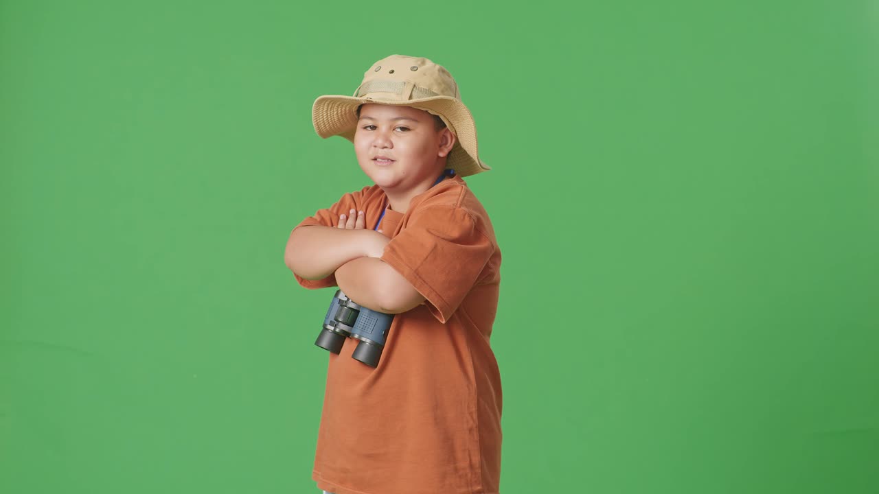 niño turista asiático con un sombrero y binoculares cruzando los brazos y sonriendo a la cámara mientras está de pie en el fondo de la pantalla verde. niño investigador examina algo, aventura de turismo de viaje, vista lateral