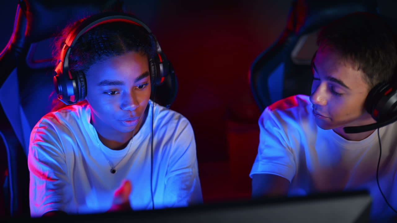White boy and black girl teens in headsets discussing while playing video games in video game club with blue and red illumination. Keyboard and mouse with illumination