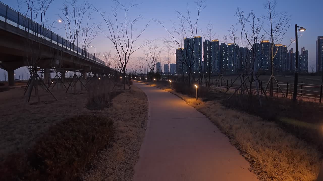 Lit Paved Pathway At Dusk Alongside Road And Bridge in Incheon, South Korea. City Buildings In Background. wide pullback shot