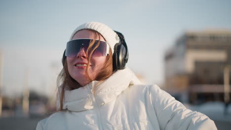 free spirit smiling joyfully under sunlight while grooving in large headphones and sunglasses, dressed in white winter jacket with soft background blur of cityscape and snowy weather