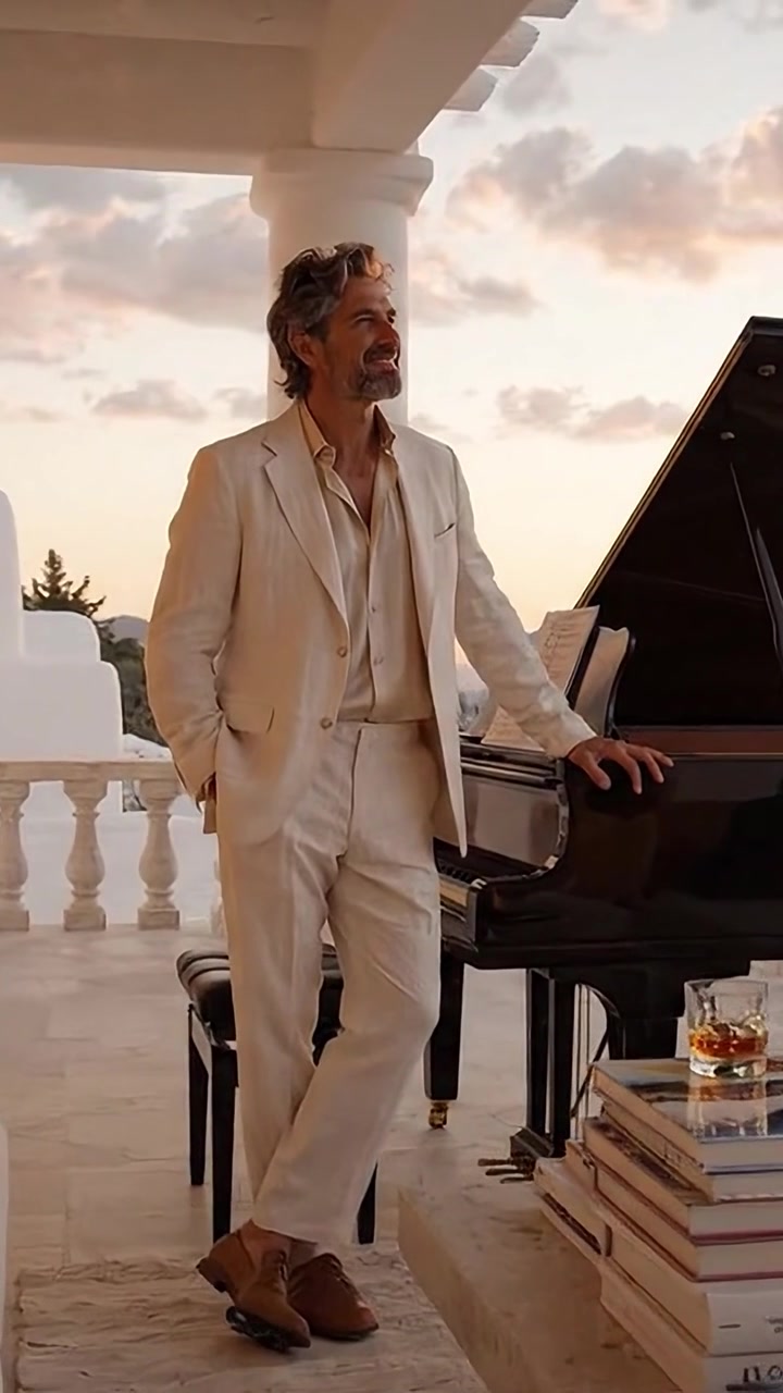 Uomo elegante in abito color crema accanto a un pianoforte a coda, che emana raffinatezza mentre si gode un tramonto sereno con un bicchiere in mano, completato da un arredamento lussuoso.