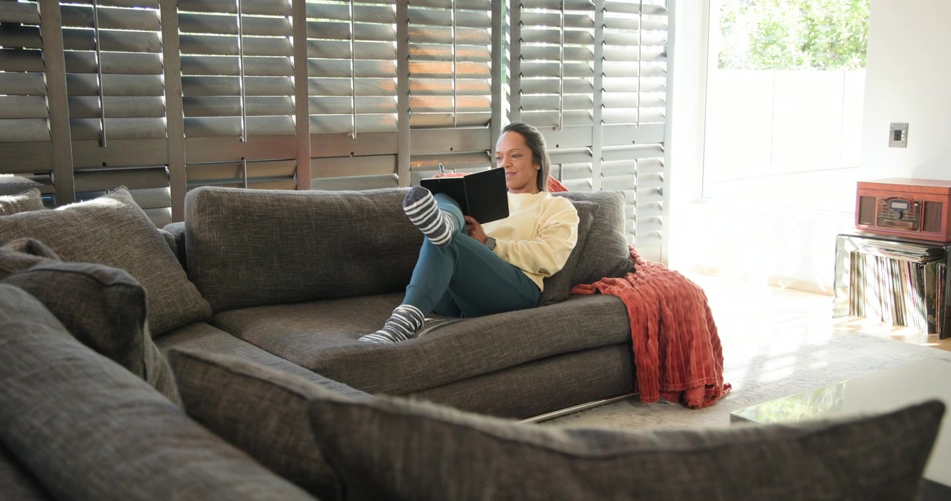 Moden afroamerikansk kvinde sidder til højre på en grå sofa og læser på en sort tablet, slapper af, med plads til tekst.