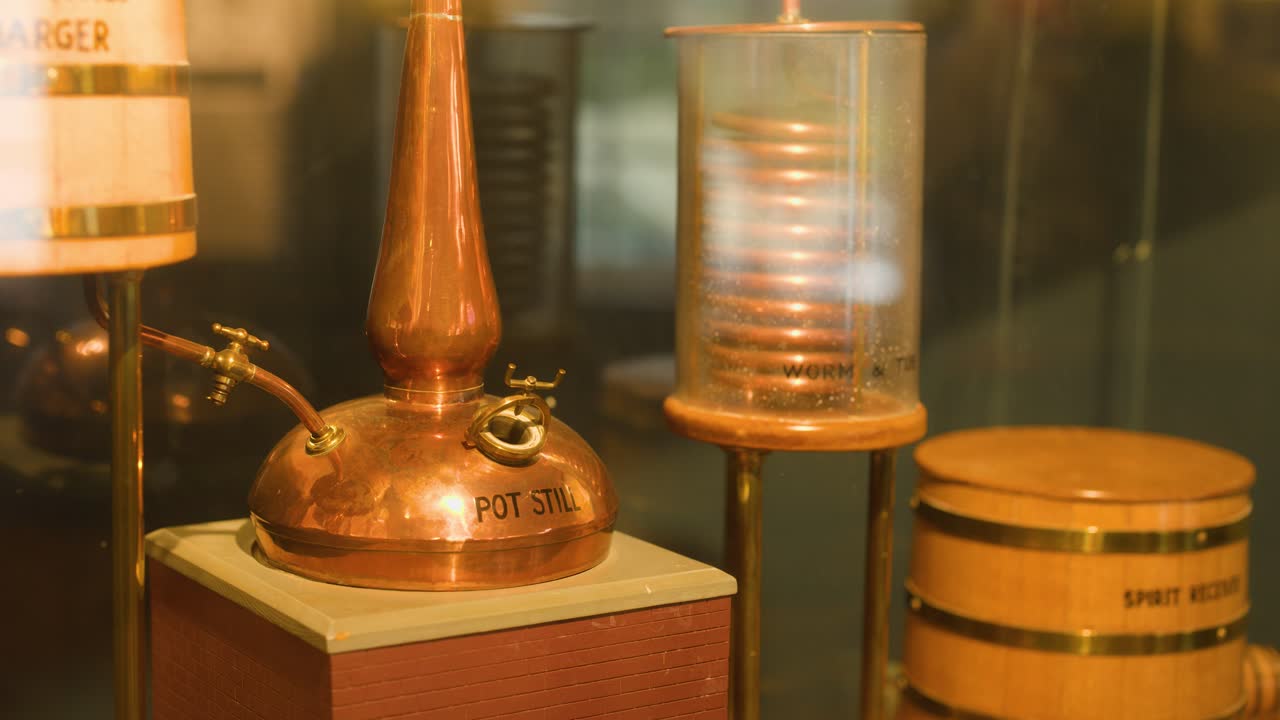 Static close-up of copper pot still, worm tub, and barrels under warm indoor lighting