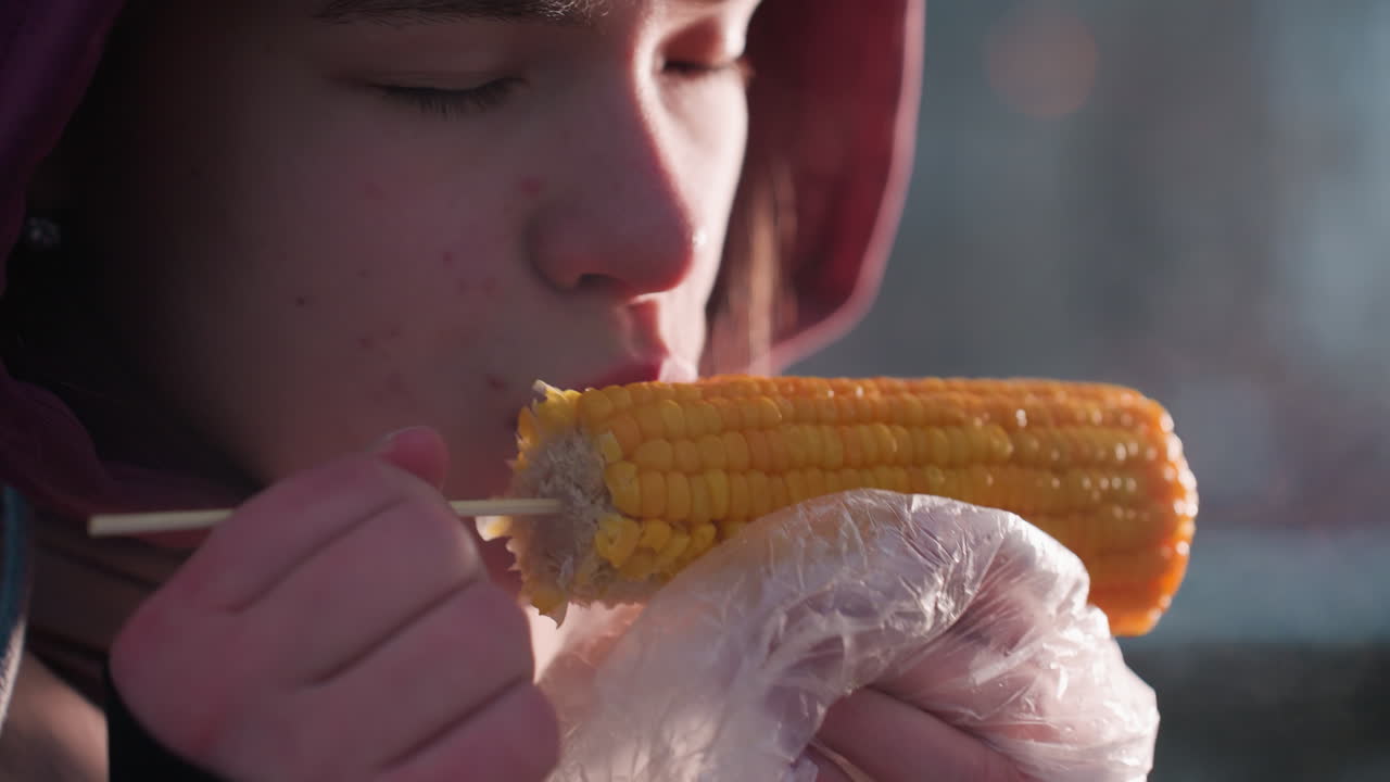 primo piano di una donna che mangia mais con vapore caldo che si alza da esso, mostrando calore e consistenza fresca, momento di cibo in un parco invernale all'aperto, involucro di plastica sul mais