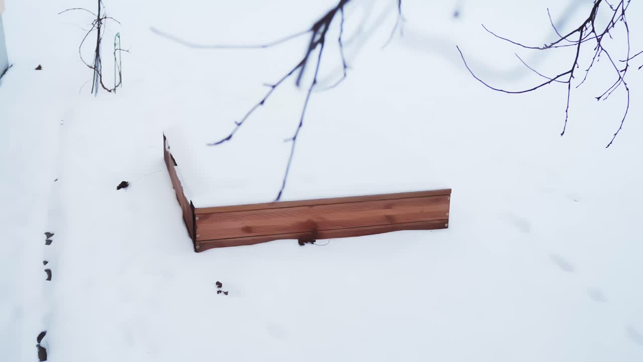 Top-down view of a wooden sandbox frame partially buried under fresh snow in a quiet backyard, showing winter season stillness, cold climate living, and outdoor prep