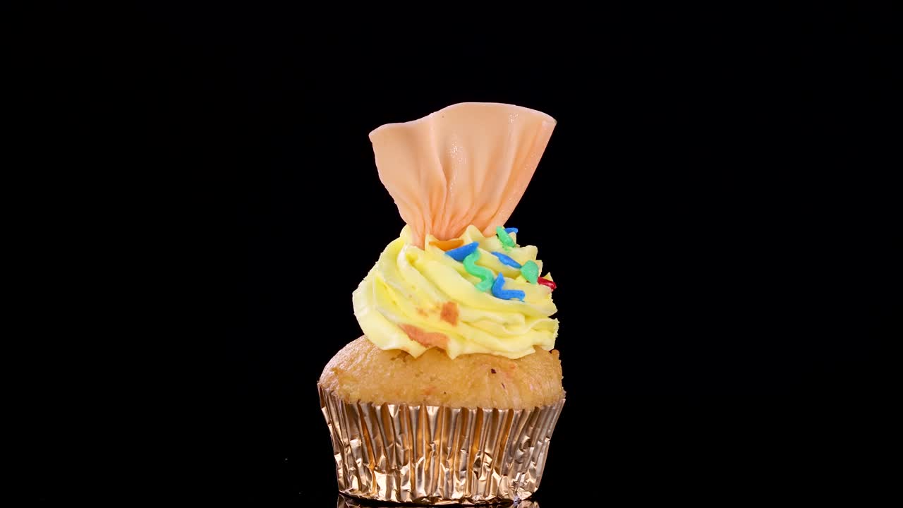 Un singolo cupcake gourmet con glassa gialla e codette colorate ruota dolcemente su uno sfondo nero senza soluzione di continuità sotto un'illuminazione da studio brillante e uniforme.