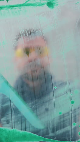 cu, low angle shot: breaded man in googles cleans green paint from glass using stripper in spacious light room closeup
