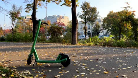 a green scooter rests on a leaf-strewn pathway in a city park, framed by autumn trees and distant buildings