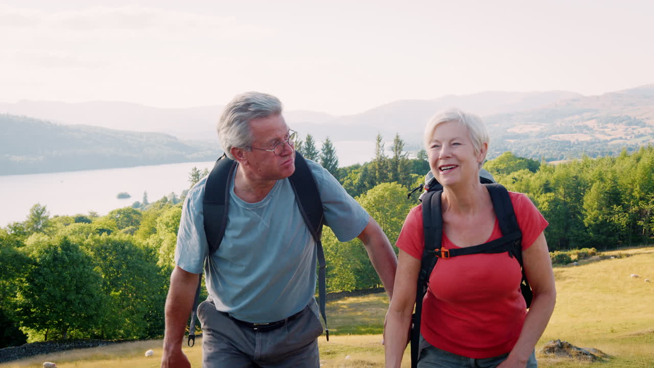 Slow Motion Shot Of Senior Couple Climbing Hill On Hike Through Countryside In Lake District UK Together
