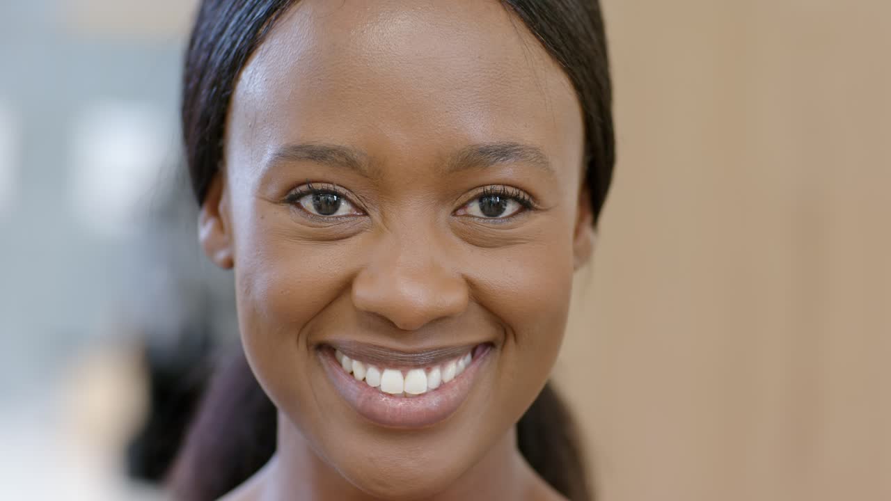 Donna africana adulta di fronte alla fotocamera in studio, sorridente e che ride per una battuta fuori campo, con indosso top in tinta neutra e orecchini a lobo