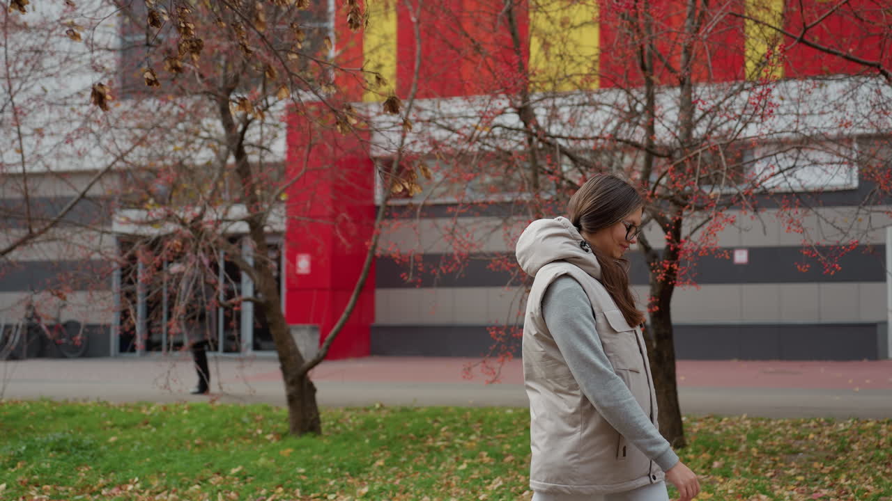 Cheerful woman walking in urban park dressed for summer with red autumn trees swaying, parked bicycles, cars on roadside, people passing by and city buildings in background