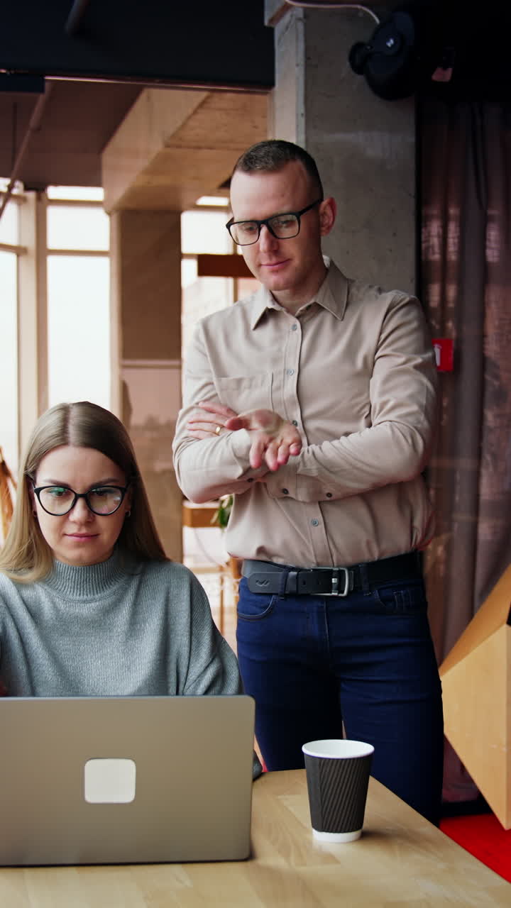 Male and female colleague in the office. Lady works at desk on her laptop. Man stands beside talking to woman. Vertical video