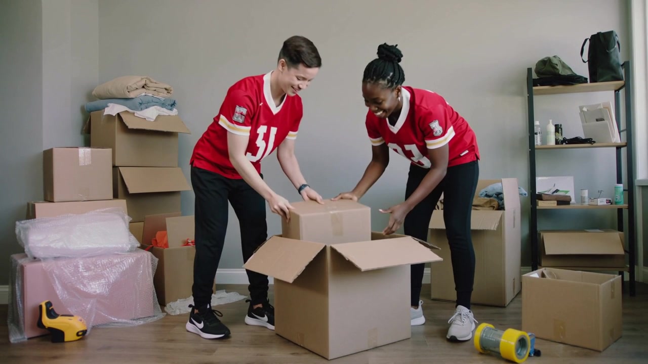 Packing two friends in red jerseys placing blue textile into large box at home, closing flaps