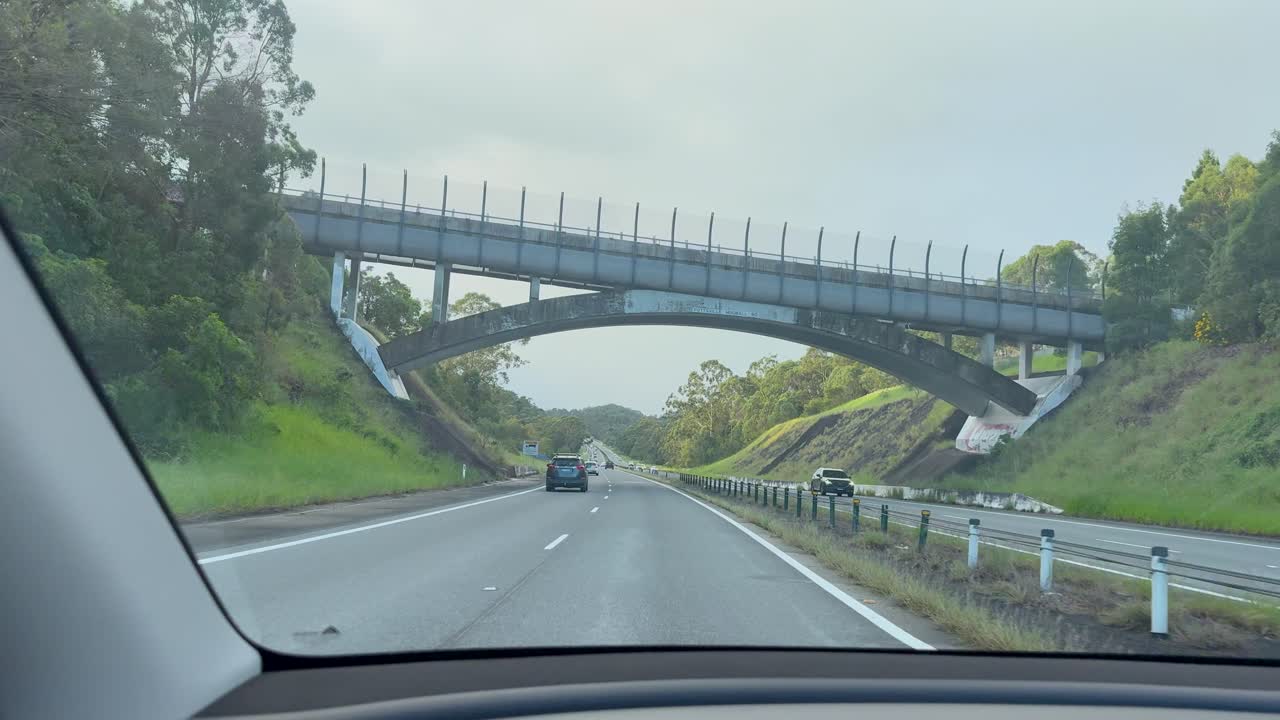 A vehicle travels along a divided highway through lush greenery, passing beneath a contemporary pedestrian bridge in daylight with steady dashboard camera movement