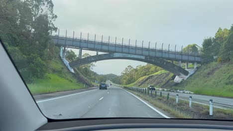 a vehicle travels along a divided highway through lush greenery, passing beneath a contemporary pedestrian bridge in daylight with steady dashboard camera movement