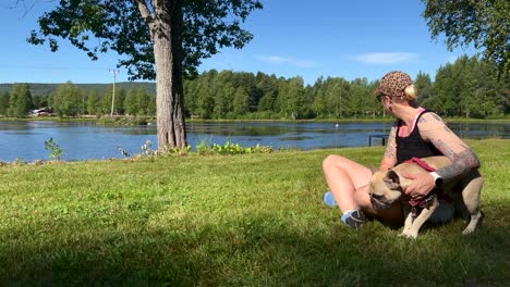 a woman sitting in the green grass petting her french bulldog in front of a lake on a sunny summer day