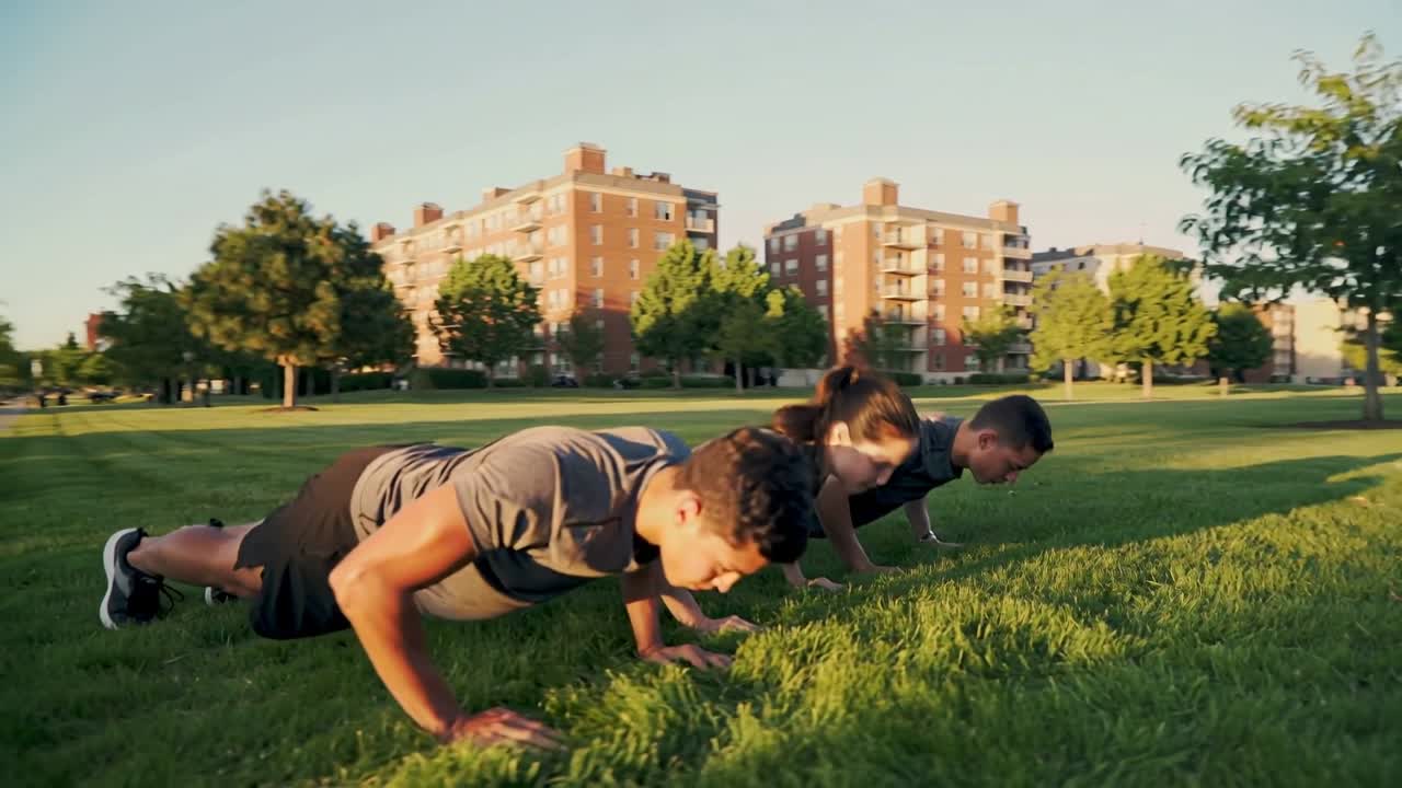 Responding to warm light, three adults doing synced push-ups on riverside lawn, wearing gray shirts