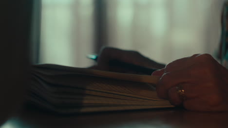 close-up-on-woman-senior-old-hands-holding-page-of-a-book-journal-while-holding-a-ink-pen-inside-house-living-room-desk