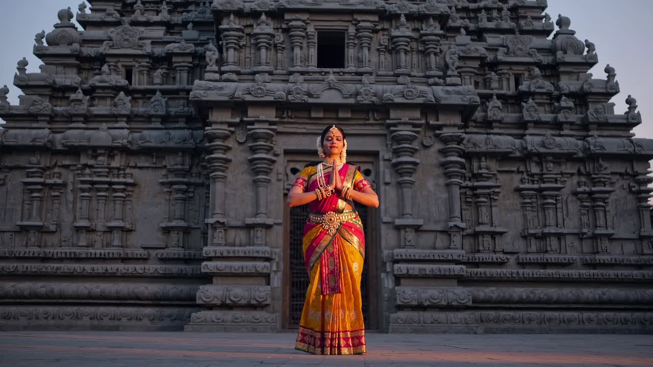 Indian classical dancer is gracefully posing in front of a temple at dawn, her colorful costume contrasting with the ancient stone architecture, creating a serene and spiritual atmosphere