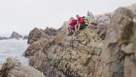 Senior-hiker-couple-with-backpacks-sitting-and-talking-on-the-rocks-while-hiking-near-sea-shore.