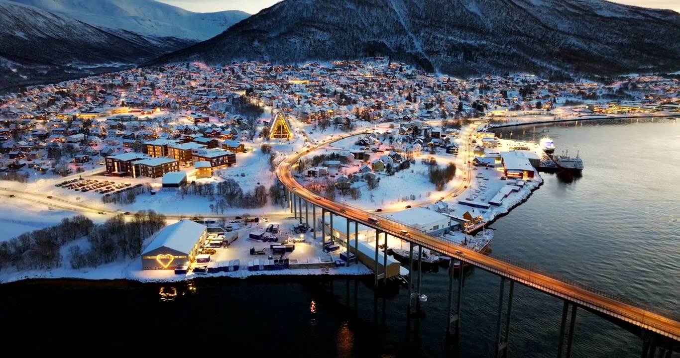 Leuchtende Straßen von Tromsø mit Verkehr über einer Brücke und schneebedeckter Stadt, Drohnenaufnahme.