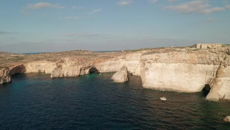 Drone-is-flying-over-clear-turquoise-waters-of-Malta's-Blue-Lagoon,-orbiting-around-a-boat-and-unveiling-rugged-rock-formations-and-coastal-caves-along-Como-Island's-shoreline