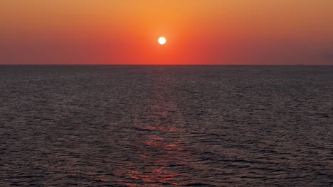 Dolly-Aéreo-Mientras-La-Bola-De-Sol-Se-Pone-En-El-Horizonte-Sobre-El-Agua-Del-Océano-Reflejando-Azul-Oscuro-Y-Rojo-Naranja