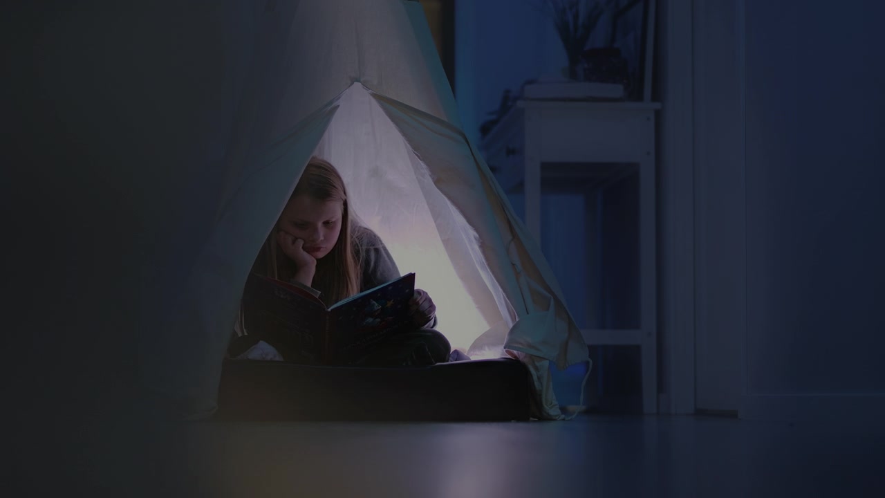 Young girl reading a book inside a cozy indoor tent at night with soft glowing light