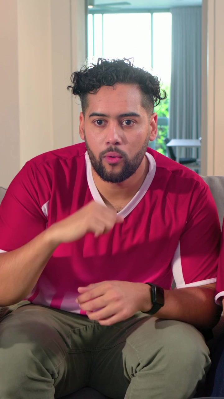 Vertical video: Clapping man reacting to television in living room, wearing red jersey, wristwatch
